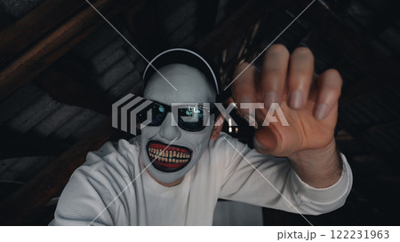 A scary masked man in a long-sleeved white sweatshirt and black cap stretches out his hand in a dimly lit attic with wooden beams. 122231963