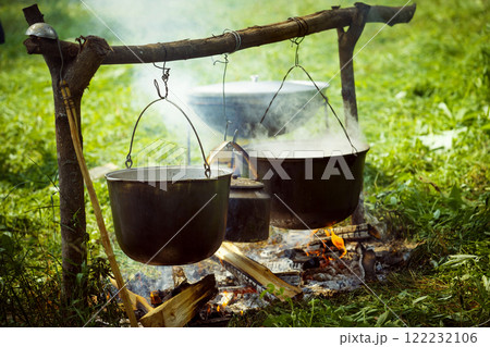 A group of cauldrons and kettle are hanged upon fire at tourist camp at green summer outdoors background. 122232106