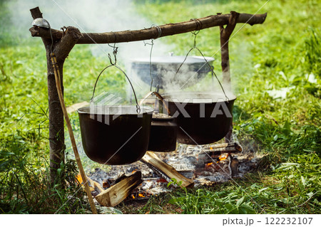A group of cauldrons and kettle are hanged upon fire at tourist camp at green summer outdoors background. A group of cauldrons and kettle are hanged upon fire at tourist camp at green summer outdoors background. 122232107