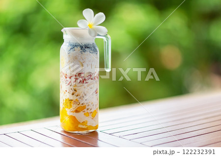 Pitcher of superfood layered smoothie with tropical fruits and berries at wooden table at green tropical blurred background with copyspace. 122232391