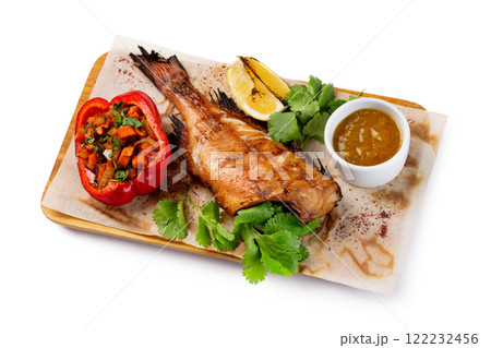 Grilled fish with no head served on paper at wooden board with parsley, pepper, lemon and sauce isolated at white background. Grilled fish with no head served on paper at wooden board with parsley, pepper, lemon and sauce isolated at white background. 122232456