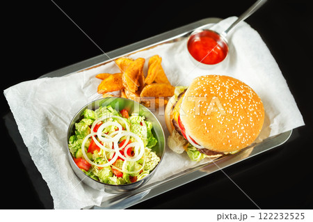 Closeup fast food set including hamburger, ketchup, salad and french fries on a tray at black background. Closeup fast food set including hamburger, ketchup, salad and french fries on a tray at black background. 122232525