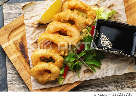 Top view image of golden crispy dried calamari rings with soy sauce served on paper at wooden table background. Top view image of golden crispy dried calamari rings with soy sauce served on paper at wooden table background. 122232730