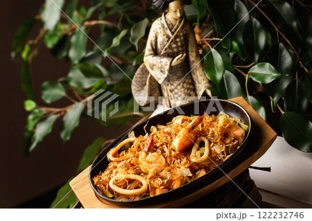 Pan of stir fried glass noodles with grilled sea food and prawns at decorated with japanese statue and green tree background. Pan of stir fried glass noodles with grilled sea food and prawns at decorated with japanese statue and green tree background. 122232746