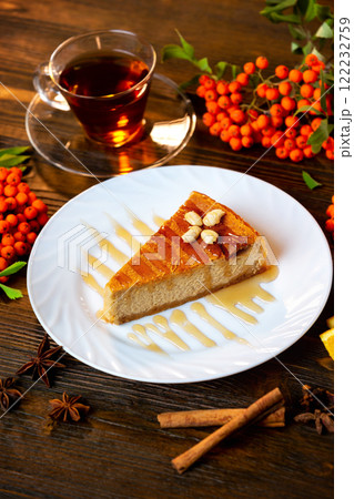 Closeup vertical image of cheesecake slice decorated with maple syrup, cup of tea and rawan branches at wooden board background. Closeup vertical image of cheesecake slice decorated with maple syrup, cup of tea and rawan branches at wooden board background. 122232759