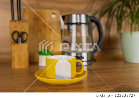 A yellow cup of tea stands on a yellow saucer and on the saucer lies a tea bag on a wooden table against the background of a teapot, a plant and kitchen utensils. The concept of tea drinking, break A yellow cup of tea stands on a yellow saucer and on the saucer lies a tea bag on a wooden table against the background of a teapot, a plant and kitchen utensils. The concept of tea drinking, break 122235727