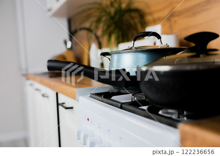 Close-up of a black frying pan with a transparent lid and a silicone handle and a blue saucepan with a transparent lid and silicone handles against the background of a modern wooden-white kitchen out 122236656
