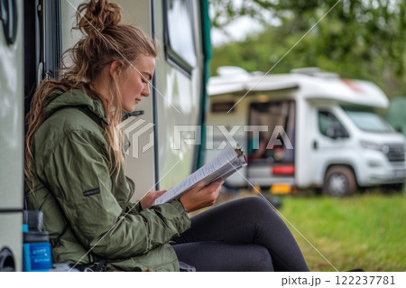 A young woman enjoys nature while reading outside her camper. Surrounded by lush greenery, she finds inspiration in the peaceful setting. Generative AI. 122237781