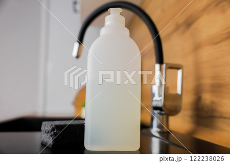Close-up of a white bottle of dishwashing soap, a black dishwashing sponge lying next to it against the background of a black granite sink and a flexible high faucet and a white wooden modern kitchen 122238026
