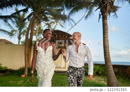 Black person in white dress holds hand with LGBTQ partner in suit at tropical wedding. Smiling couple under palm trees. Inclusive marriage, modern love diversity, joy. 122241383