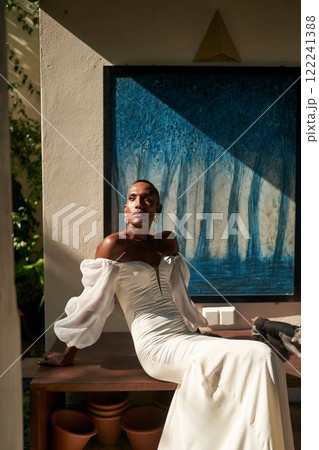 Bride in white dress sits in sunlit villa courtyard. Ambiguous gender fluid ethnic black person in wedding attire. LGBTQ wedding scene. Elegant and serene vibe. Contemporary and inclusive celebration. 122241388