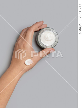 Female hand holding opened face cream jar over grey top view, negative space. Daily skincare 122241714