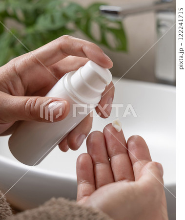 Woman in brown bathrobe pumping cream on finger and holding a cosmetic bottle in bathroom 122241715