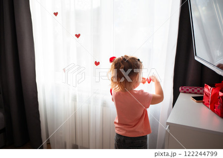 Back view of little Caucasian girl decorate the curtains on the window with red paper hearts. A celebration for Valentine's Day and Mother's Day. Copy space Back view of little Caucasian girl decorate the curtains on the window with red paper hearts. A celebration for Valentine's Day and Mother's Day. Copy space 122244799