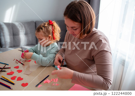 Caucasian adult mother and her little cute daughter cut out paper hearts sitting at a desk. Family is preparing for the celebration of Valentine's Day 122244800