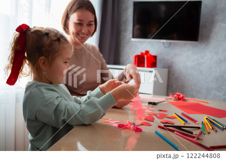 Side view of Caucasian woman kindergarten teacher teaches a little cute girl how to cut hearts out of red paper for Valentine's Day Side view of Caucasian woman kindergarten teacher teaches a little cute girl how to cut hearts out of red paper for Valentine's Day 122244808