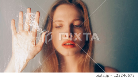 Female isolation. Sadness art. Sensual disturbed woman standing behind wrinkled polyethylene film on blur pastel pink blue color gradient glow background. Female isolation. Sadness art. Sensual disturbed woman standing behind wrinkled polyethylene film on blur pastel pink blue color gradient glow background. 122246675