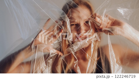 Lonely woman. Depressive mood. Disturbed concerned girl trapped behind wrinkled transparent polyethylene film isolated on light background. 122246719