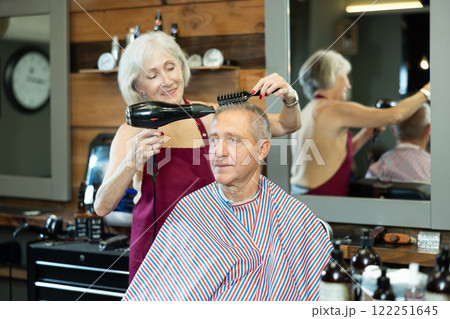 Female barber using hairdryer to style hair to elderly client in barbershop Female barber using hairdryer to style hair to elderly client in barbershop 122251645