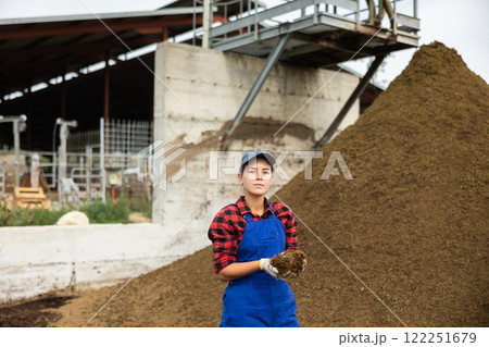 Female farmer with shovel poses against pile of dried and crushed cow dung in the backyard of farm 122251679