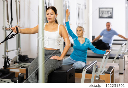 Calm smiling young woman in sportswear pulling hand in band from Pilates reformer machine during workout in small studio 122251801