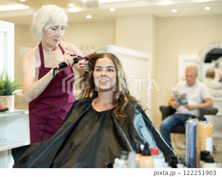 Elderly female hairstylist using curling wands to add volume for hair of young girl 122251903