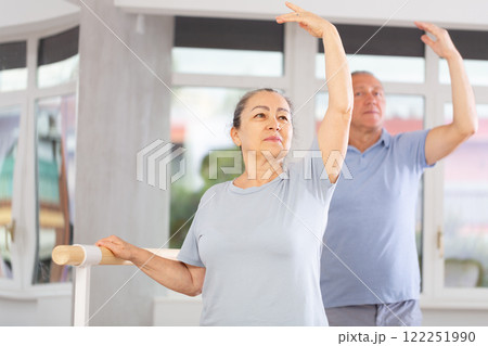 Elderly women and men practice at ballet standing in fourth position at barre in choreography studio Elderly women and men practice at ballet standing in fourth position at barre in choreography studio 122251990