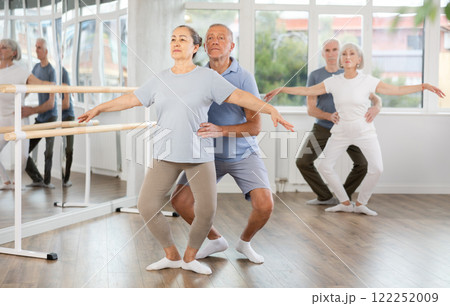 Diligent couple of elderly modern ballet dancers practicing demi plie ballet poses with support in hall with ballet bar Diligent couple of elderly modern ballet dancers practicing demi plie ballet poses with support in hall with ballet bar 122252009
