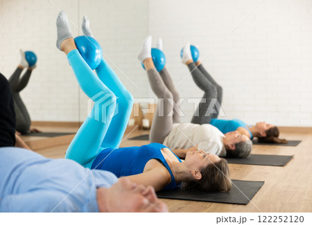 Class of people of different ages holding balls between legs and pushing press during Pilates training in studio 122252120