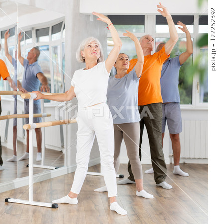 Smiling mature woman in sports clothing with group active aged people training ballet dance movements, standing in fourth position at barre in dance salon 122252392