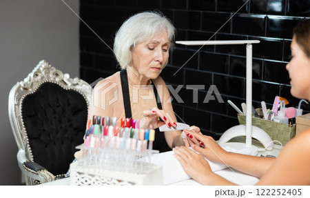 Friendly elderly woman working in beauty salon as manicure master shaping the nails of young female client with nail file 122252405