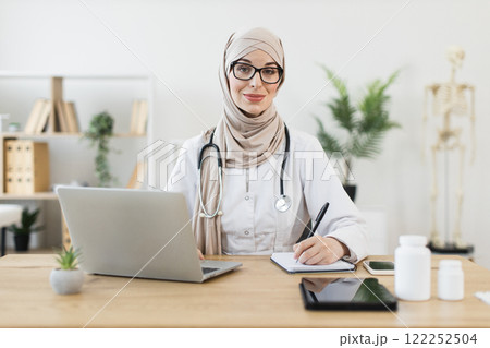 Young Muslim female doctor using laptop for online consultation, writing notes. Medical professional working in modern office. Confident adult woman in hijab providing healthcare remotely. 122252504