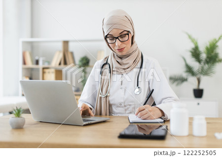 Young Muslim female doctor wearing hijab conducting video call with patient, writing prescription. Professional medical consultation in modern office. Technology enhancing communication in healthcare. Young Muslim female doctor wearing hijab conducting video call with patient, writing prescription. Professional medical consultation in modern office. Technology enhancing communication in healthcare. 122252505
