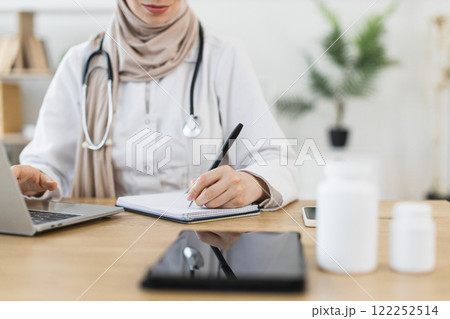 Muslim female doctor in white coat having video call with patient, using laptop, writing prescription. Middle-aged professional in medical environment. Muslim female doctor in white coat having video call with patient, using laptop, writing prescription. Middle-aged professional in medical environment. 122252514