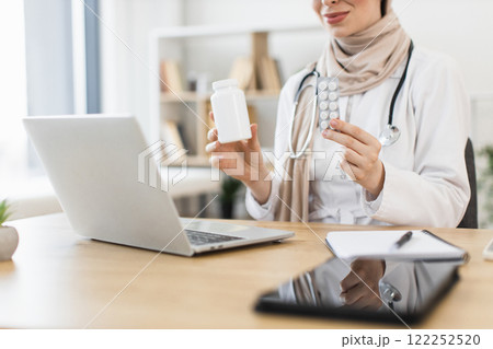 Muslim female doctor demonstrates medication during virtual consultation with patient. Middle-Eastern woman uses laptop to offer pills as treatment, exemplifying telemedicine. Muslim female doctor demonstrates medication during virtual consultation with patient. Middle-Eastern woman uses laptop to offer pills as treatment, exemplifying telemedicine. 122252520