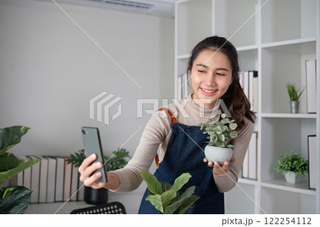 A young Asian woman uses her phone to take photos and enjoys planting plants in her garden to create a pleasant atmosphere in the house. 122254112