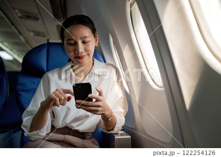 Young Asian woman uses mobile phone to check news information Sitting near the window in business class, airplane class during flight, travel and business concept 122254126