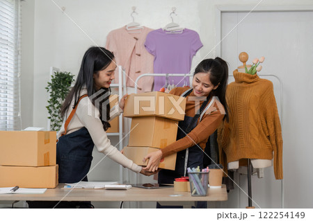 Two women entrepreneurs joyfully exchanging packaged shirt orders while managing their online boutique 122254149