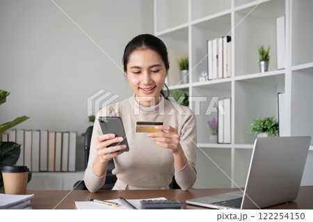 Beautiful young woman uses laptop to shop online, buys things and pays with credit card through phone online banking app. 122254150