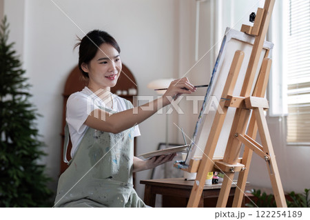 Young Asian female artist is intently painting acrylic paints on canvas in a painting studio. 122254189