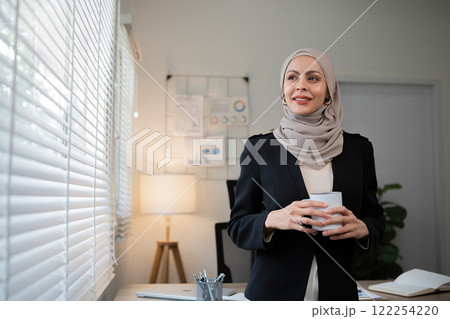 Confident Muslim businesswoman standing with coffee mug in front of desk in office during morning time 122254220