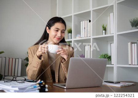 Young Asian business woman sits and works using a laptop in a modern office decorated with shady green plants. 122254321