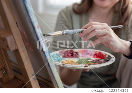 Young Asian artist woman holding a palette of colorful paints is intently painting acrylic paints on canvas in a painting studio. 122254391