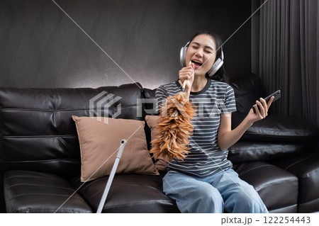 Cheerful and happy young woman singing and dancing while cleaning the living room, holding house cleaning equipment and wearing headphones, singing happily. 122254443