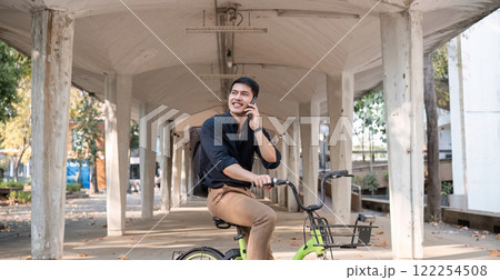 A young Asian businessman rides a bicycle to work. Standing outside the office talking on the phone and talking about business. Concept of reducing energy and reducing air pollution. A young Asian businessman rides a bicycle to work. Standing outside the office talking on the phone and talking about business. Concept of reducing energy and reducing air pollution. 122254508