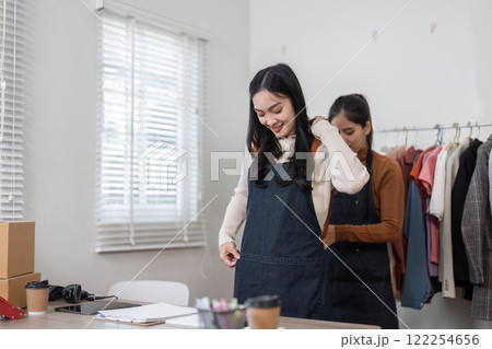 Women entrepreneurs trying on stylish aprons while preparing for their online shirt business in a modern boutique Women entrepreneurs trying on stylish aprons while preparing for their online shirt business in a modern boutique 122254656