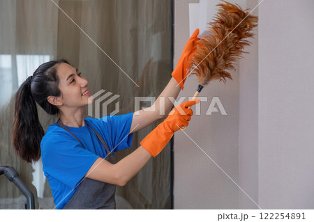 A beautiful young housekeeper does housework wearing orange rubber and uses a duster to clean up the items in the room. 122254891
