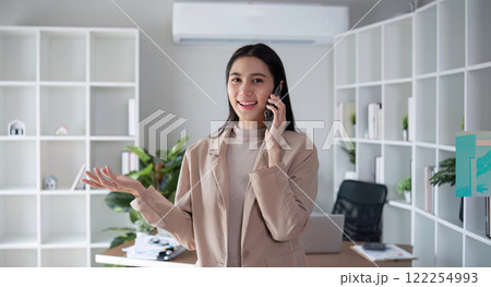Young Asian business woman sits on the phone in an online business meeting using a laptop in a modern home office decorated with shady green plants. 122254993