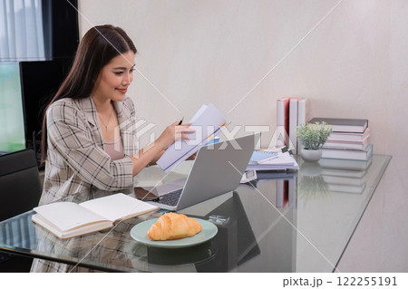 A middle-aged woman works from home at a dining room table with snacks and piles of documents on it. 122255191