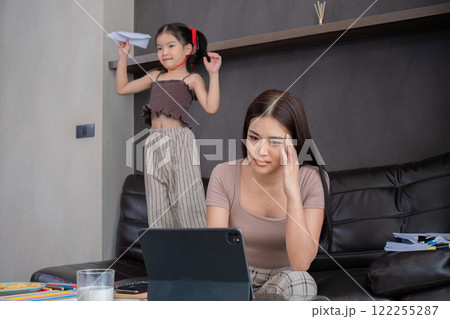 Stressed business mother sitting from home, taking notes and looking after her laptop with her cute daughter playing next to her. 122255287
